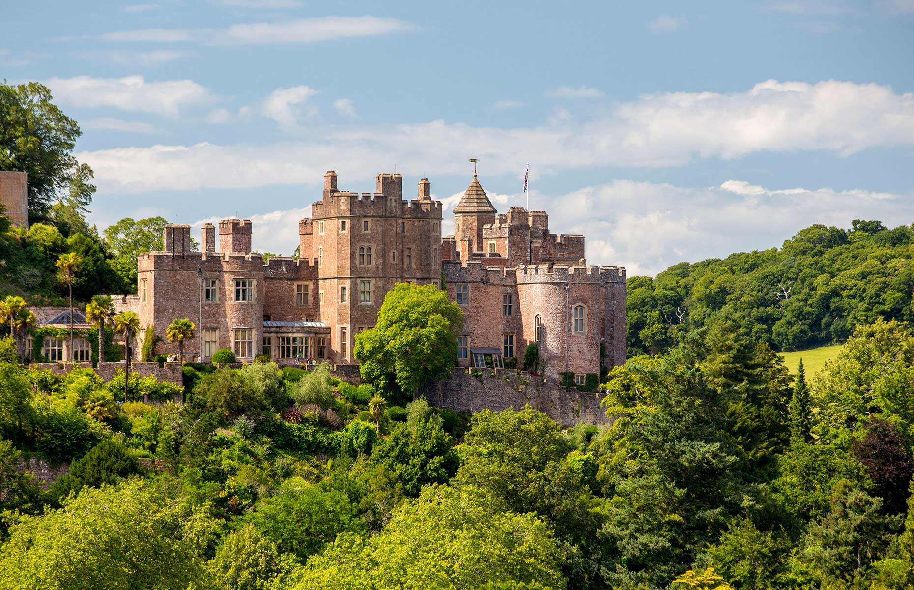 Dunster Castle viewed from a distance in summer with the red brick contrasting against the green trees (Andrew Duke/Alamy Stock Photo)