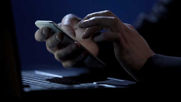 Man holding a mobile phone near a computer. (Image: Shutterstock)